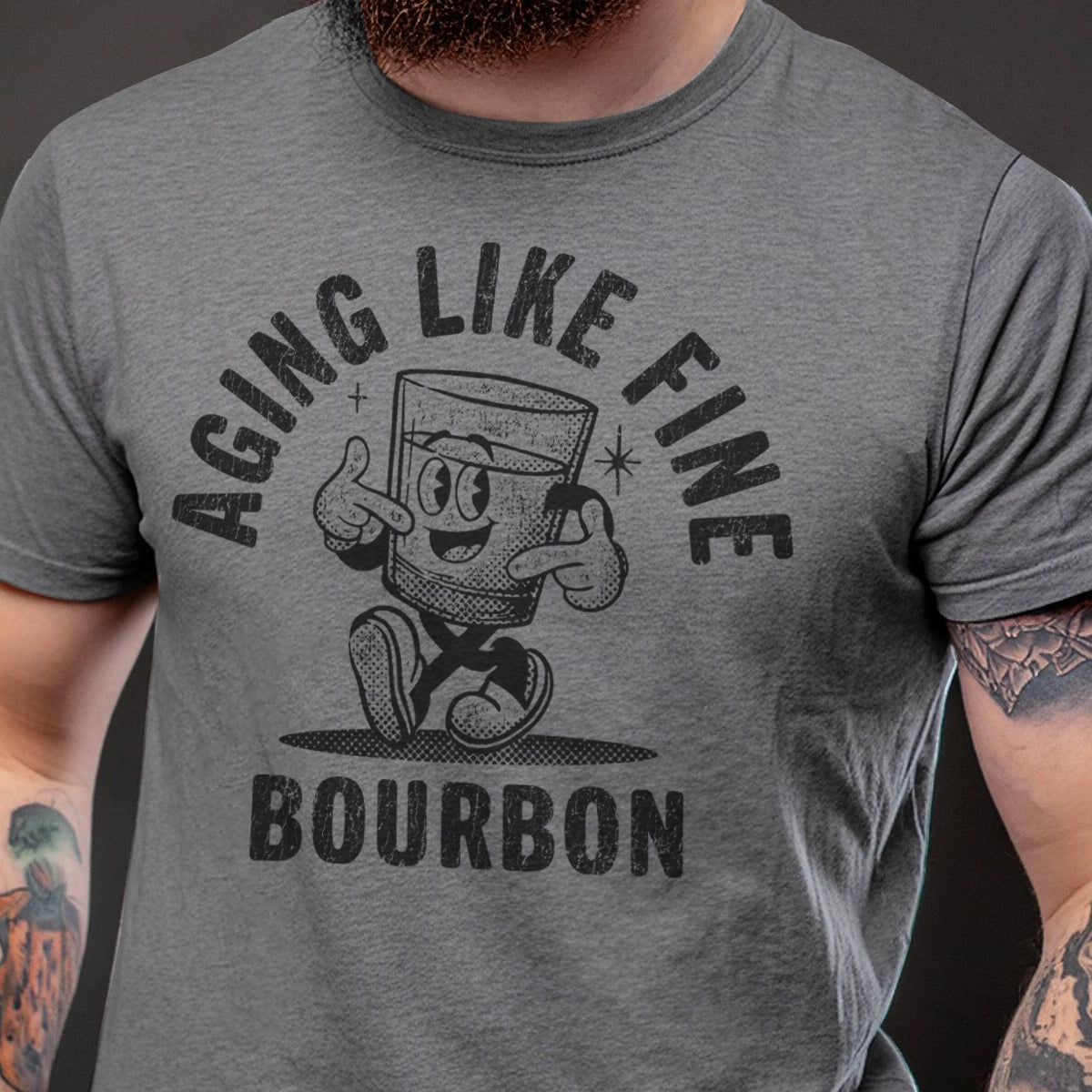 Gray t-shirt with a graphic of a cartoon glass of bourbon and text 'Aging Like Fine Bourbon' on a dark background.