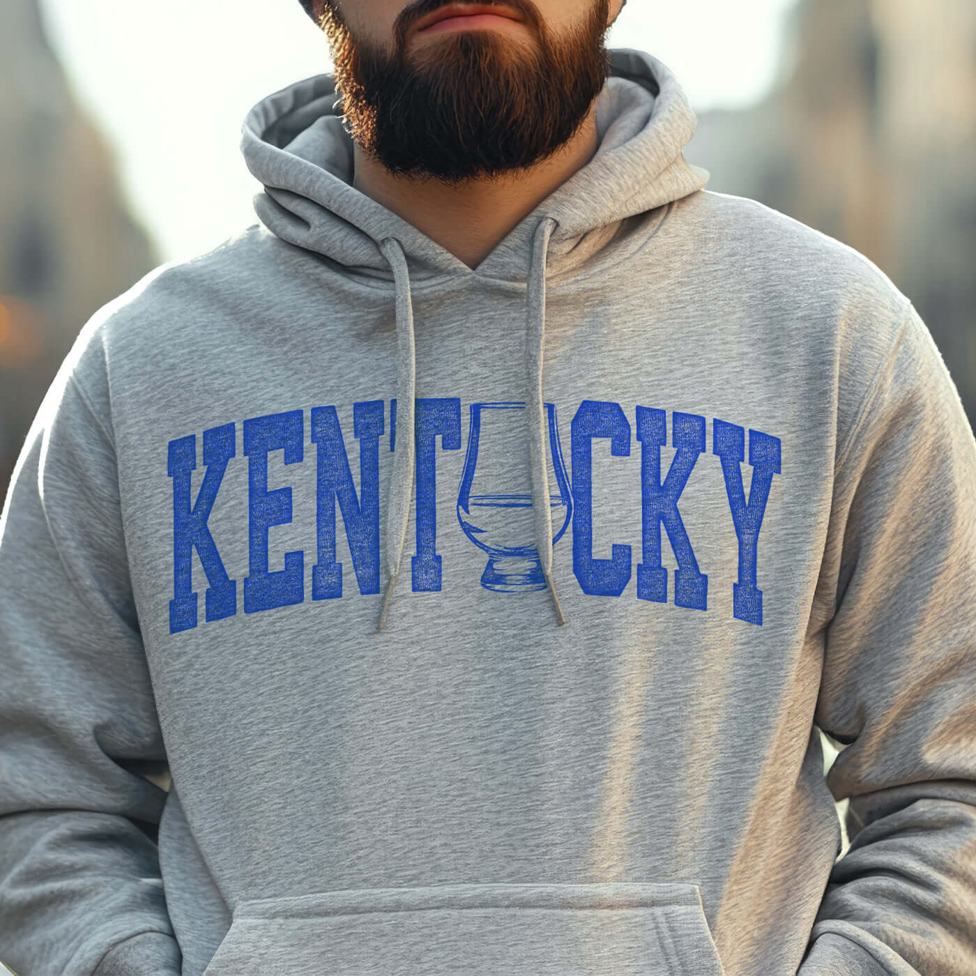 Man wearing a heather grey Kentucky bourbon hoodie with blue lettering outdoors.