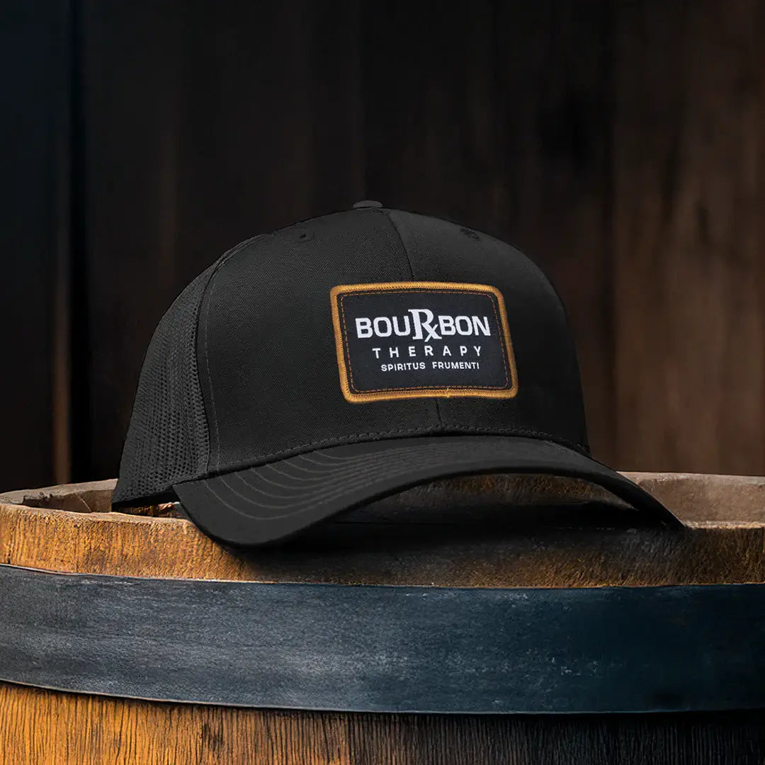 Black Richardson 112 cap with 'Bourbon Therapy' patch on a wooden barrel against a dark background