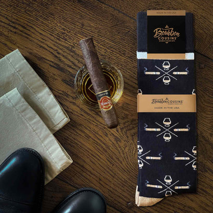 Bourbon+Cigar Socks in black with cigar and whiskey glass pattern, styled with a cigar and whiskey on wooden table.