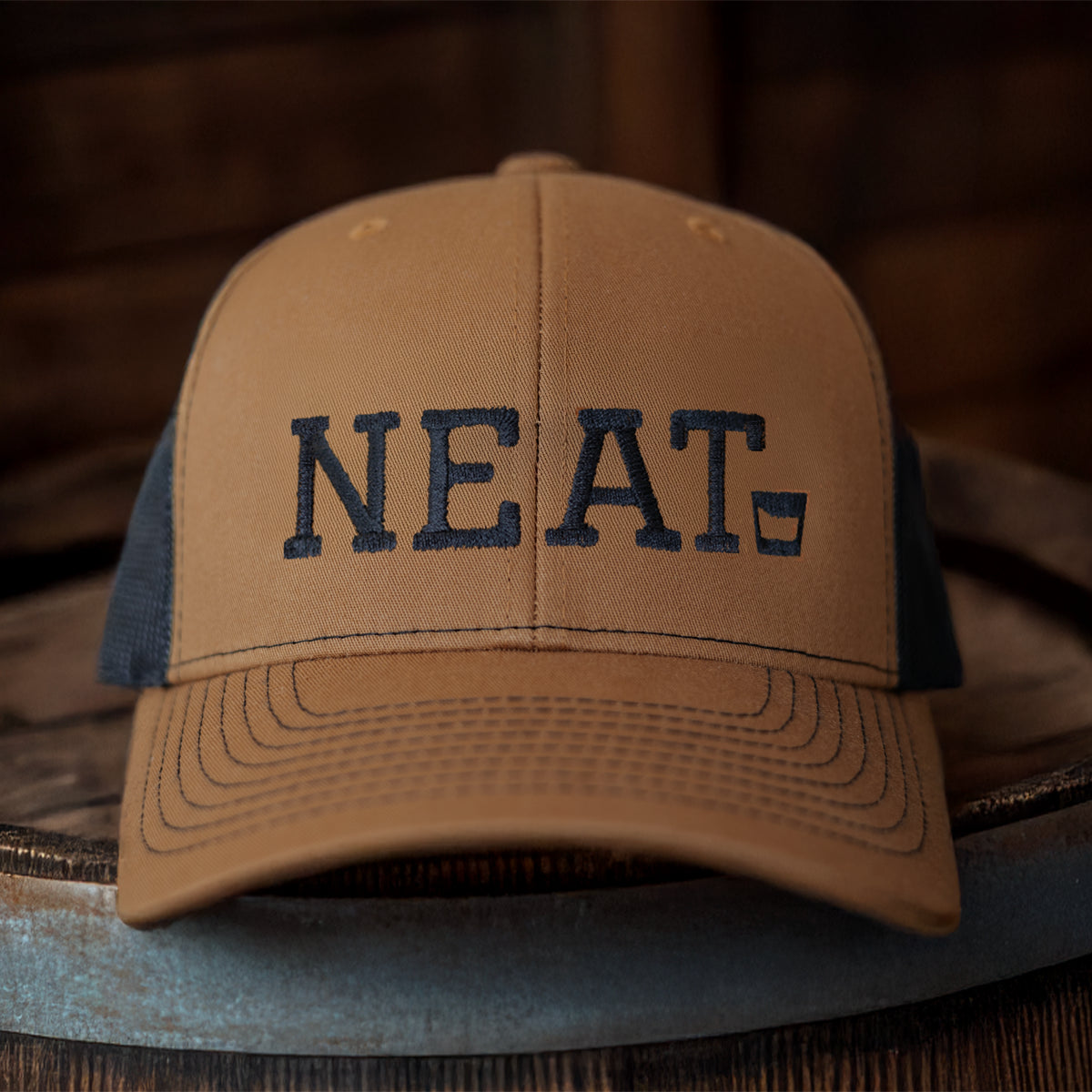 caramel and black Richardson 112 hat sitting on a bourbon barrel with an embroidered whiskey graphic that reads "Neat" 