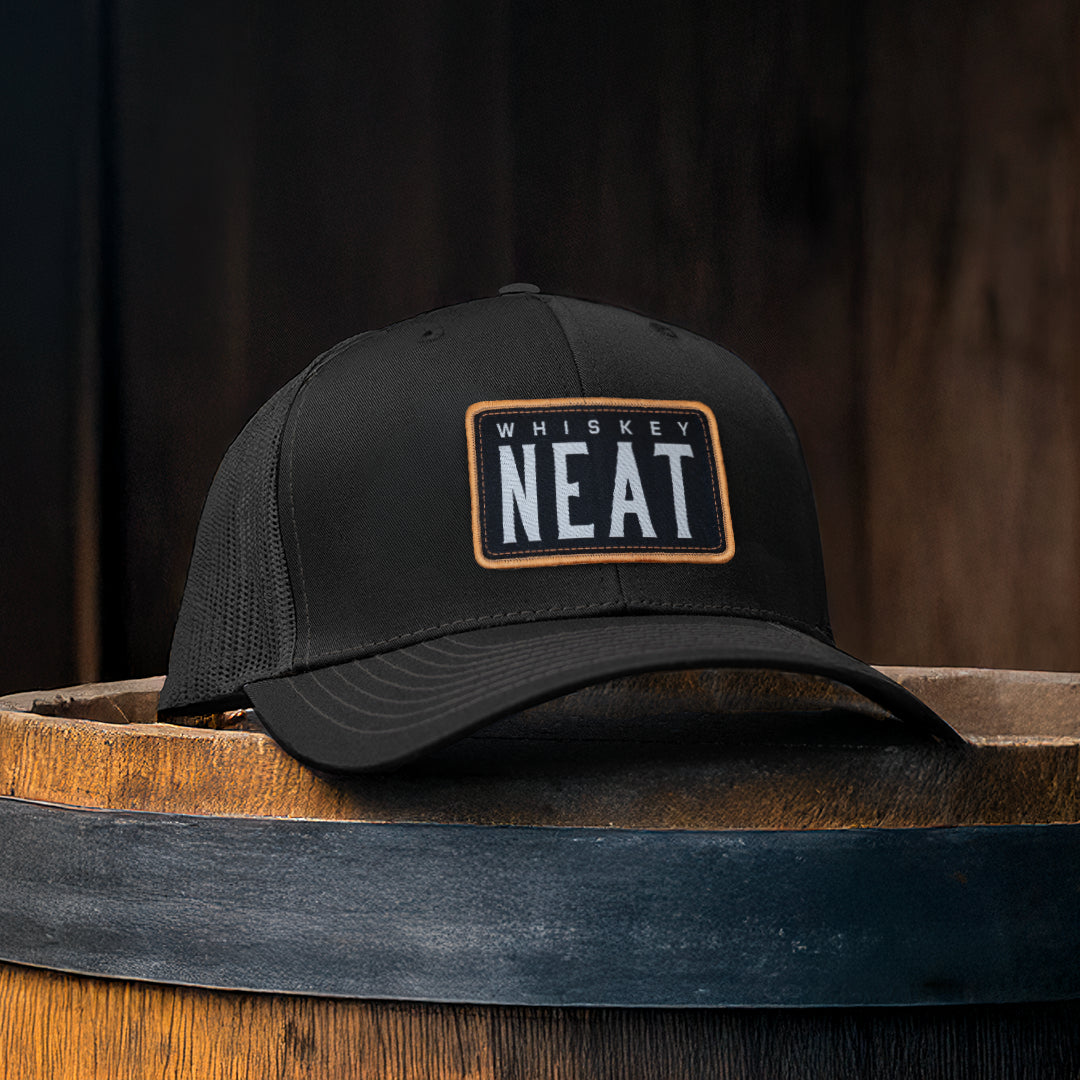 NEAT Whiskey Hat - Black with structured front panel featuring a woven patch on wooden barrel backdrop.